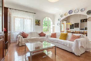 A living room with two white sofas, a glass coffee table, and a dining area with wooden chairs.