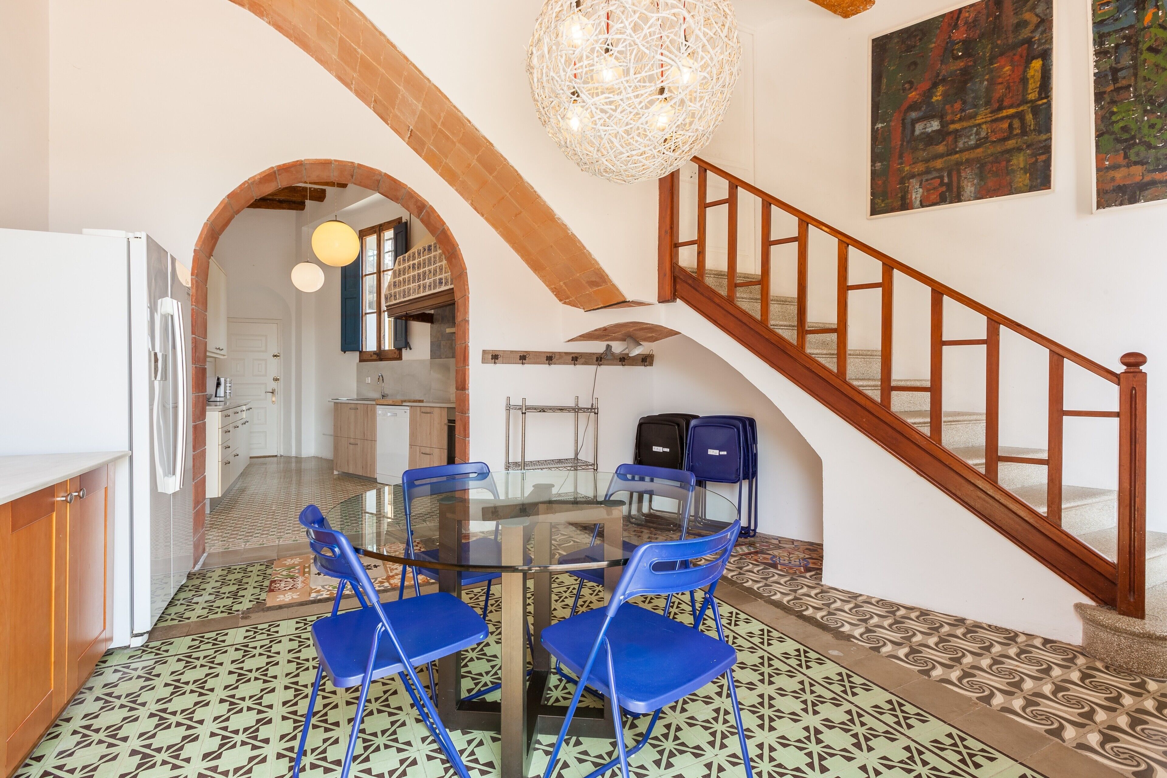 Een eethoek met een glazen tafel en blauwe stoelen, een keuken met een witte koelkast, en een trap met houten leuningen.