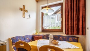 Ein Essbereich mit Tisch, Stühlen, einem Kreuz an der Wand und einem Fenster mit Vorhängen.