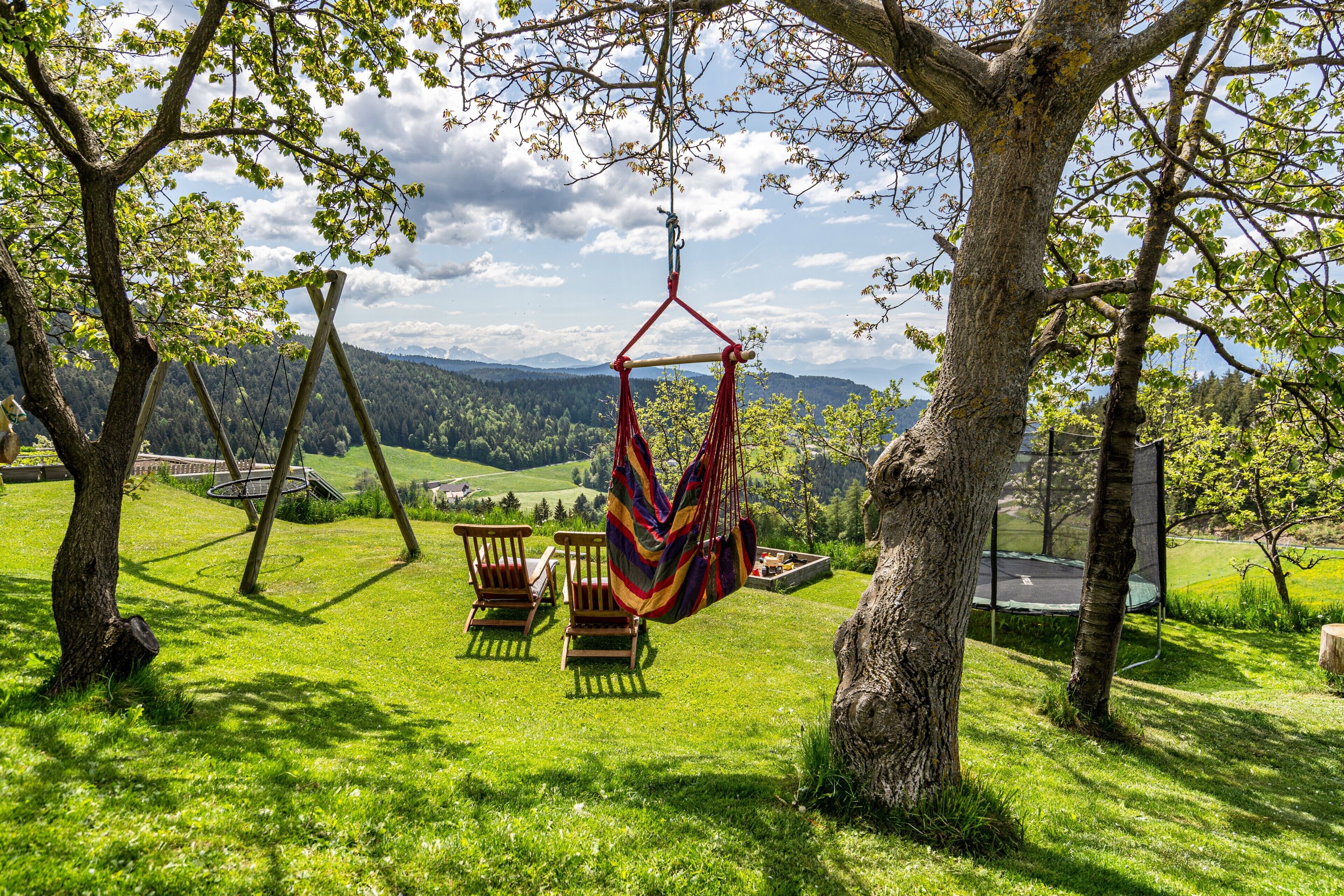 Una hamaca colgada de un árbol, sillas de madera y un espacio con césped con vistas a colinas distantes.