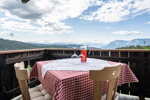 Ein Tisch mit kariertem Tuch, zwei Stühle und eine Glaskaraffe auf einem Balkon mit Bergblick.