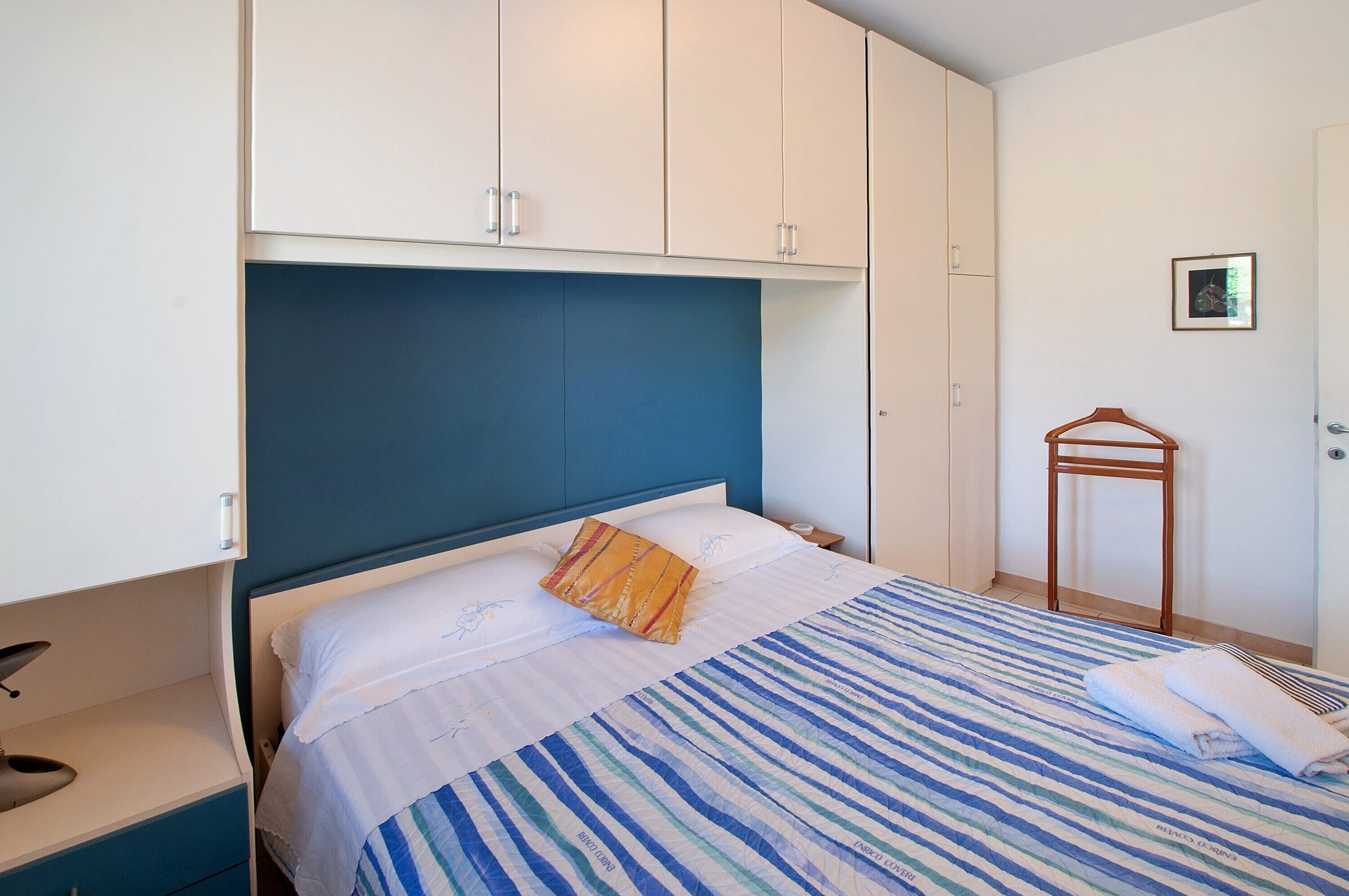 Ein ordentlich bezogenes Bett mit gestreifter Bettwäsche, ein Nachttisch mit einer Lampe und ein Holzkleiderhaken an der Wand.