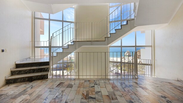 Un interior espacioso con una escalera de mármol, ventanas amplias y vista al mar.