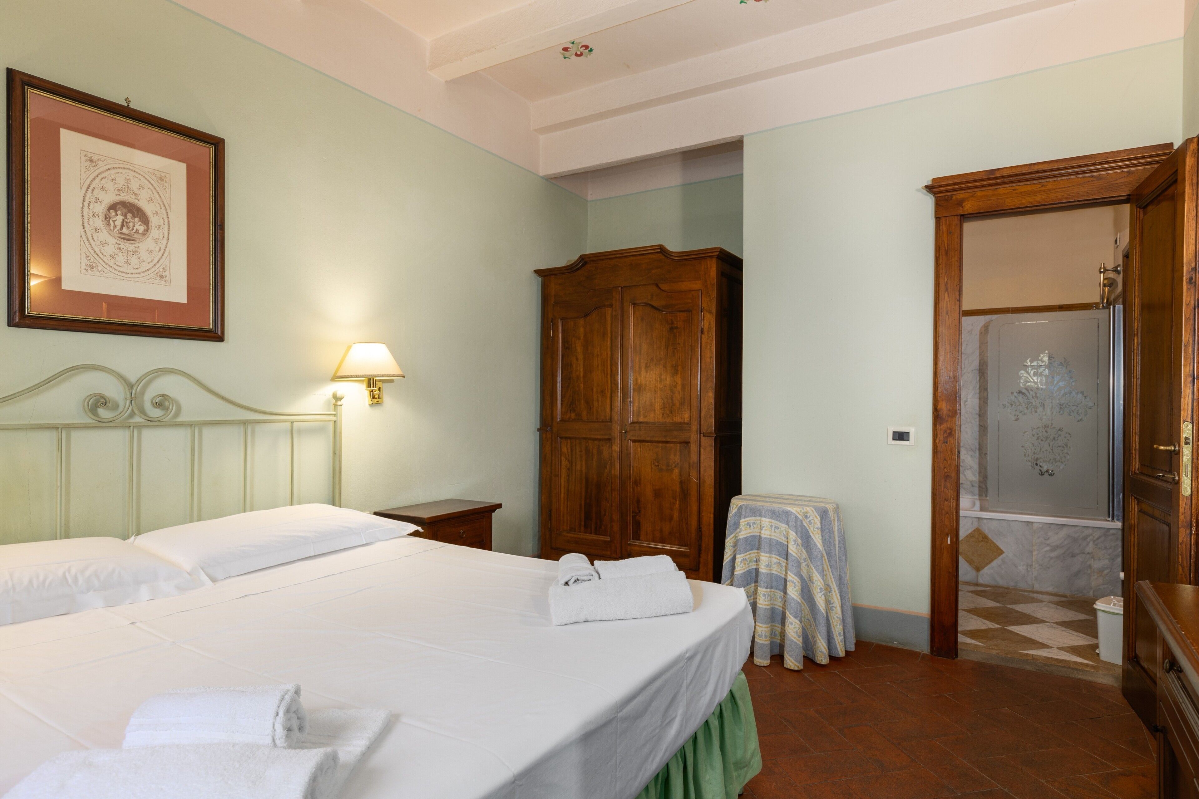 A hotel room with a bed, a wooden wardrobe, a small table with a tablecloth, and a bathroom with a shower.