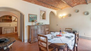 A dining area with a table set for four, a kitchen in the background, and a rustic ceiling. - Villa 'Villa del Fattore' with Shared Pool, Private Terrace & Wi-Fi (Montespertoli)