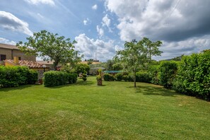 Un jardin bien entretenu, agrémenté de divers arbustes et arbres, un ciel dégagé et, au loin, un bâtiment dont le toit est recouvert de tuiles.