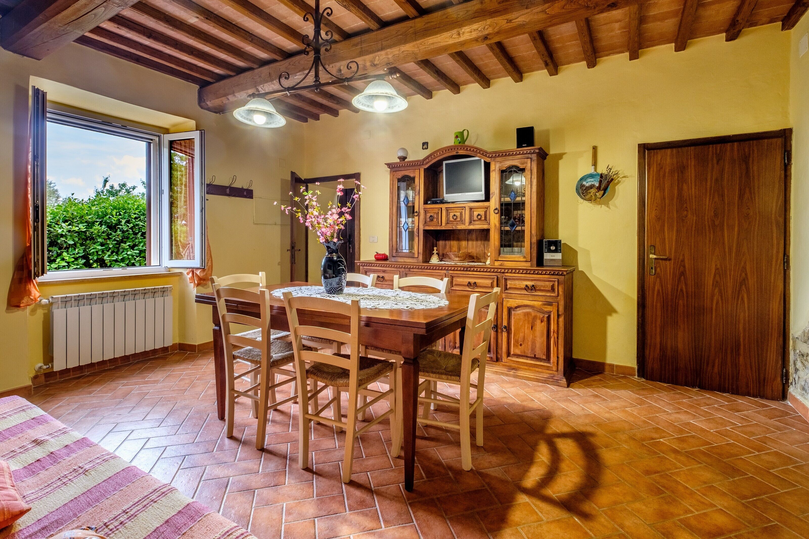 A dining area with a wooden table and chairs, a window with curtains, a radiator, and a wooden cabinet with a television.