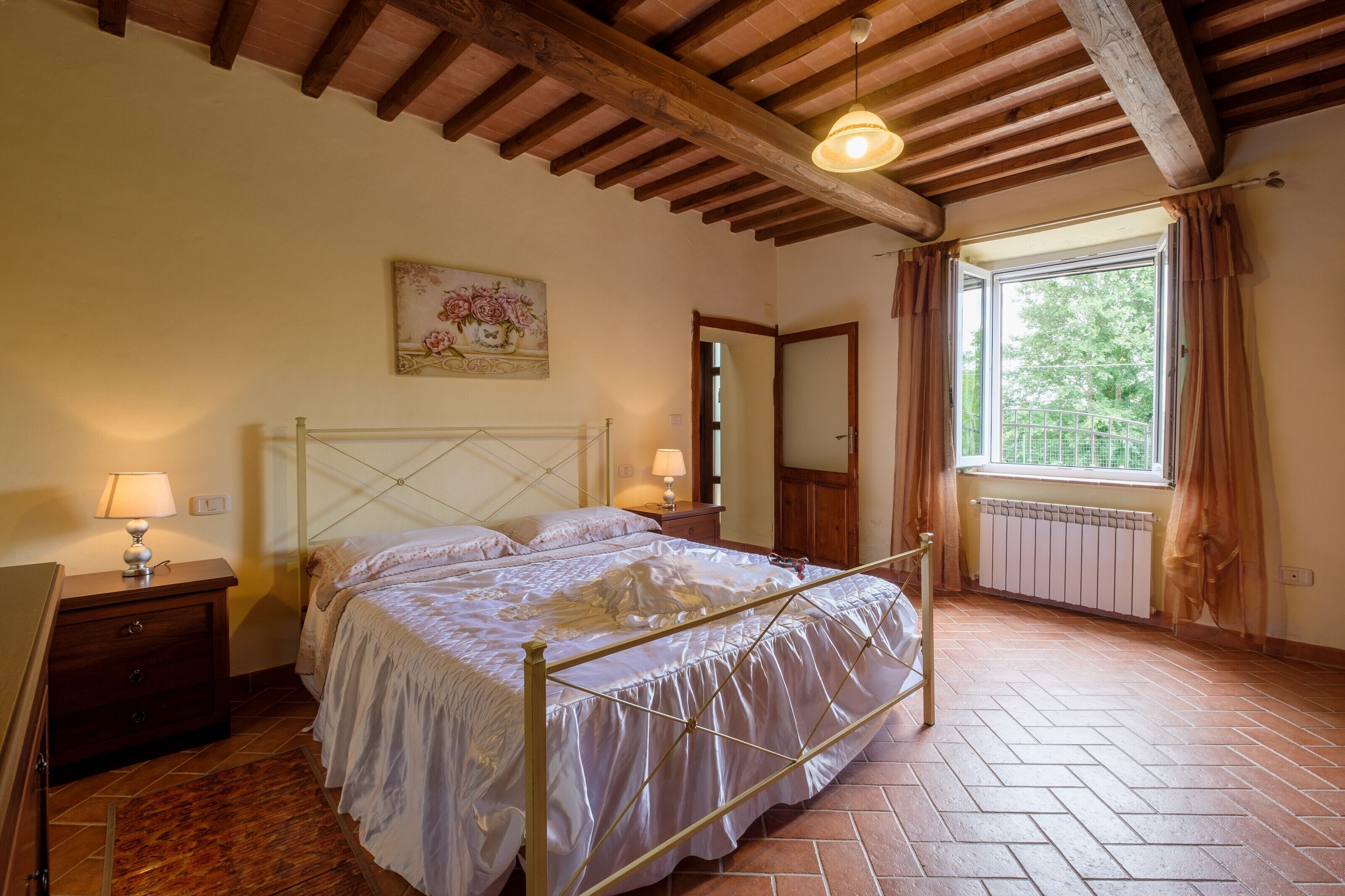 A bedroom with a bed, wooden ceiling beams, a window with curtains, a radiator, and a painting on the wall.