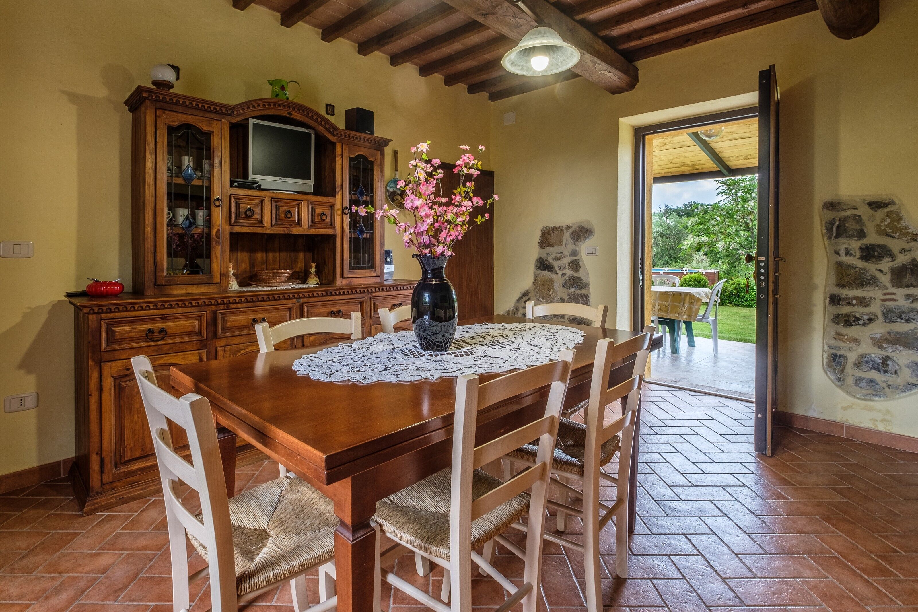A wooden dining table with chairs, a vase with flowers, a television on a cabinet, and a stone wall.