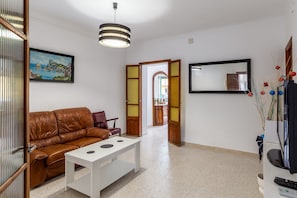 Uma sala de estar com um sofá de couro marrom, uma mesa branca, luminária pendente, um quadro emoldurado, um espelho e uma televisão.