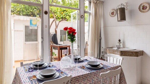 A dining area with a table set for four, a vase with red flowers, and a view of a garden through large windows. - Holiday Home 'Cas Llimoner' close to the Sea with Terrace & Wi-Fi (Palma)