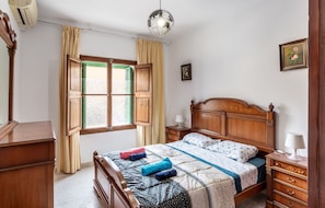 Quarto com cama de madeira, mesa de cabeceira, cômoda, janela com cortinas e ar-condicionado fixado na parede.