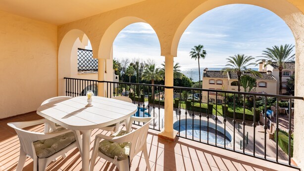 Uma varanda com mesa e cadeiras brancas, uma piscina e vista para uma área residencial com palmeiras e céu claro.