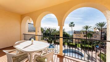 A balcony with a white table and chairs, a pool, and a view of a residential area with palm trees and a clear sky.