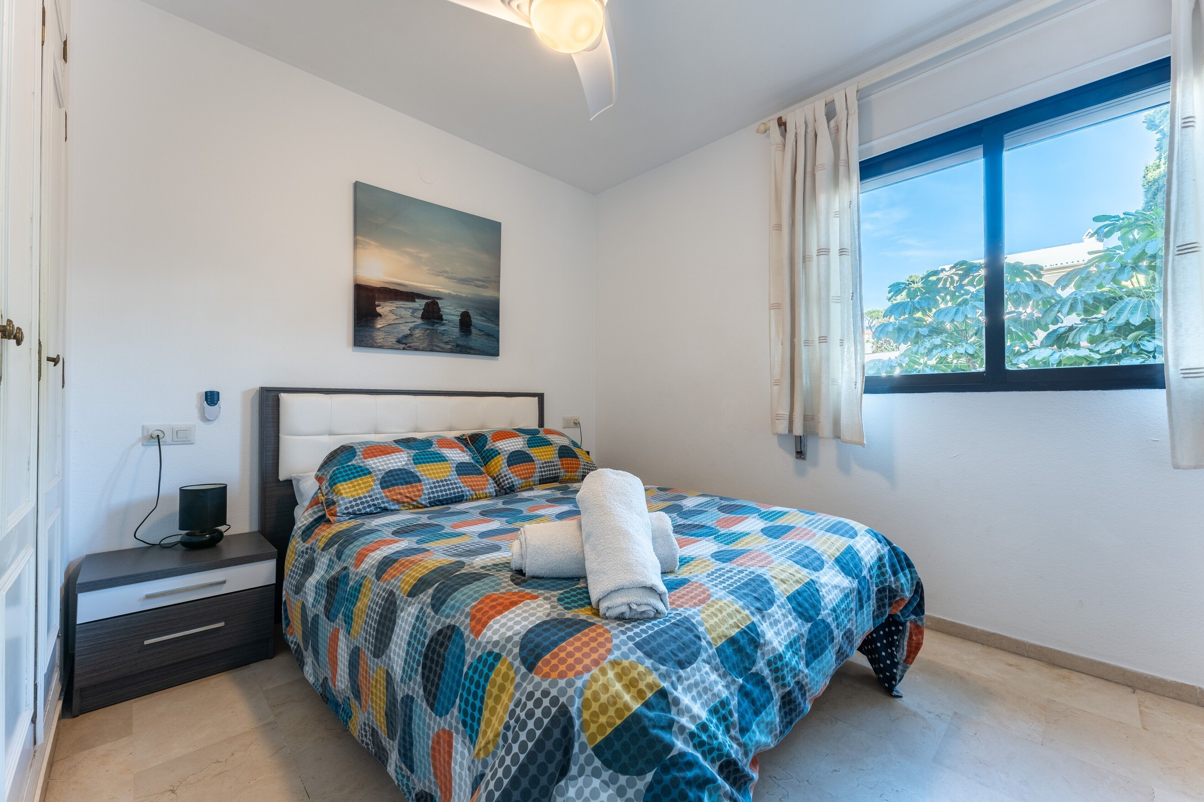 A bedroom with a bed, a nightstand, a painting, and a window with curtains.