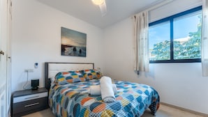 Quarto com cama, mesa de cabeceira, quadro e janela com cortinas.