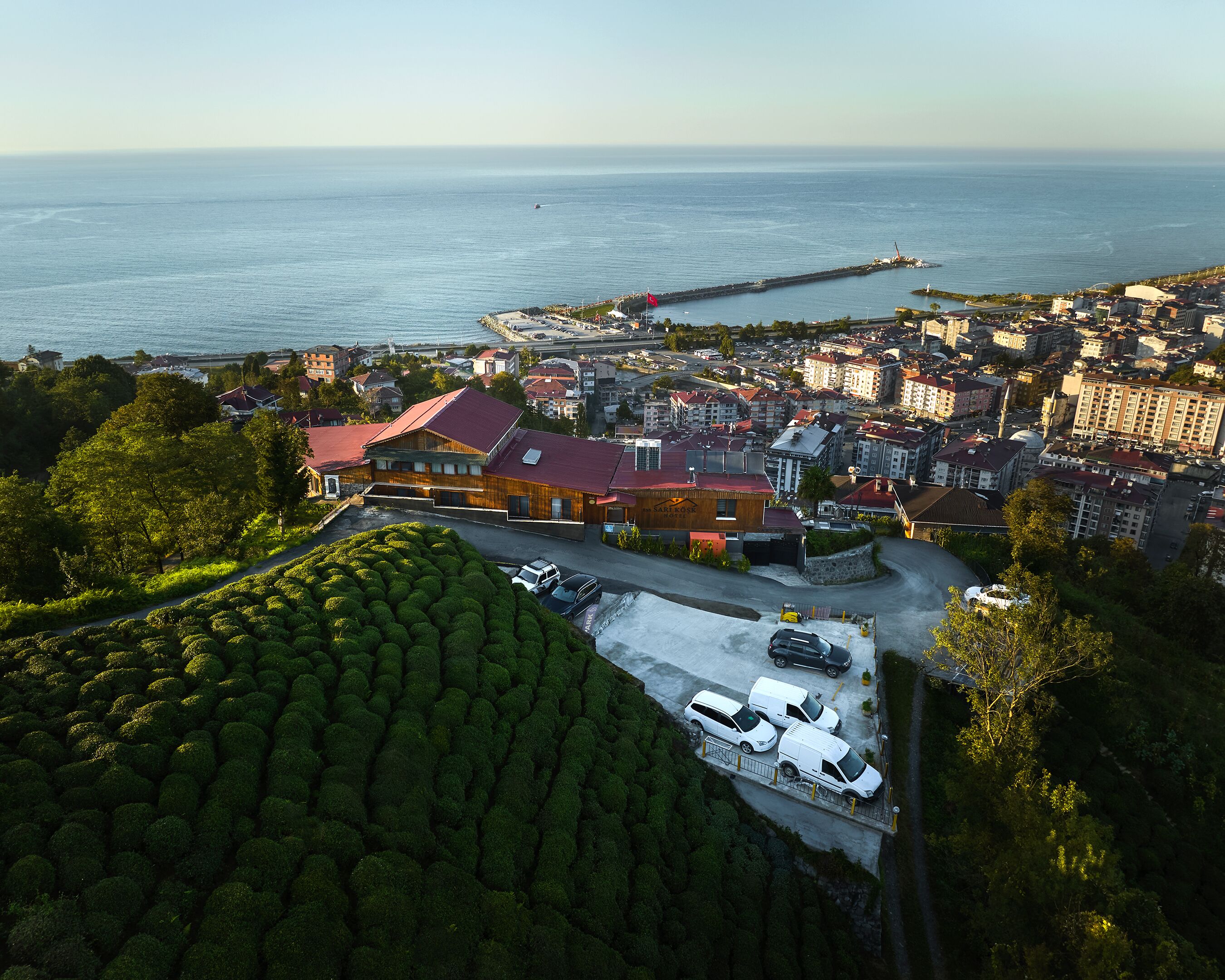 Vista aérea de un pueblo costero con un edificio destacado de techo rojo, un muelle que se adentra en el mar y un estacionamiento con varios vehículos.