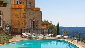 Une piscine avec des chaises longues, un bâtiment en pierre doté de balcons et une vue sur la mer.