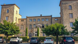 Un grand bâtiment en pierre, doté de nombreuses fenêtres et d’une tour, entouré de voitures garées et d’arbres.