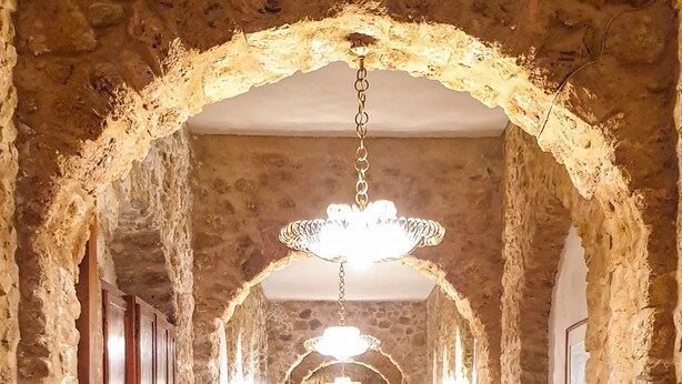 Un couloir aux murs de pierre, aux portes en bois et orné d’un lustre.