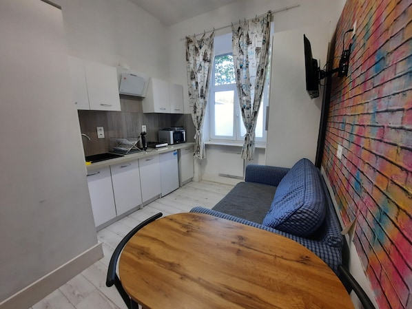 A compact kitchen with white cabinets, a microwave, and a sink. There's a wooden table and a blue sofa with a colorful brick wall. - Small apartment with Free WiFi in Krakow (Kraków)