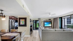 Un salon moderne avec un canapé gris, un coin repas avec des chaises en bois, une cuisine équipée d’appareils électroménagers en acier inoxydable et un téléviseur diffusant une scène de plage.