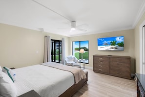 Un dormitorio con cama, ventilador de techo, televisión, cómoda y vista a una playa a través de un ventanal grande.