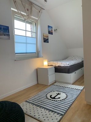 A bedroom with a bed, bedside table, lamp, window with blinds, and a rug with an anchor design.
