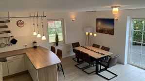 Una cocina moderna con una isla de madera, una mesa de comedor con sillas, lámparas colgantes y un ventanal con vistas a zonas verdes.