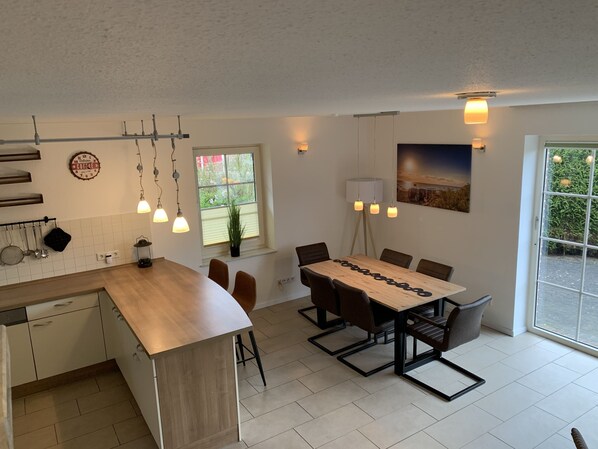 A modern kitchen with a wooden island, a dining table with chairs, pendant lights, and a large window with a view of greenery.