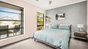 Une chambre avec une grande fenêtre, un lit recouvert d’une couvre-lit bleu clair, une table de chevet avec une lampe et un tableau accroché au mur.