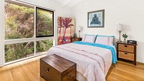 A bedroom with a large window, a bed with blue and white bedding, a wooden nightstand, a bedside table with a lamp, and a wall-mounted painting. - The Dream in Smiths Beach (Smiths Beach)