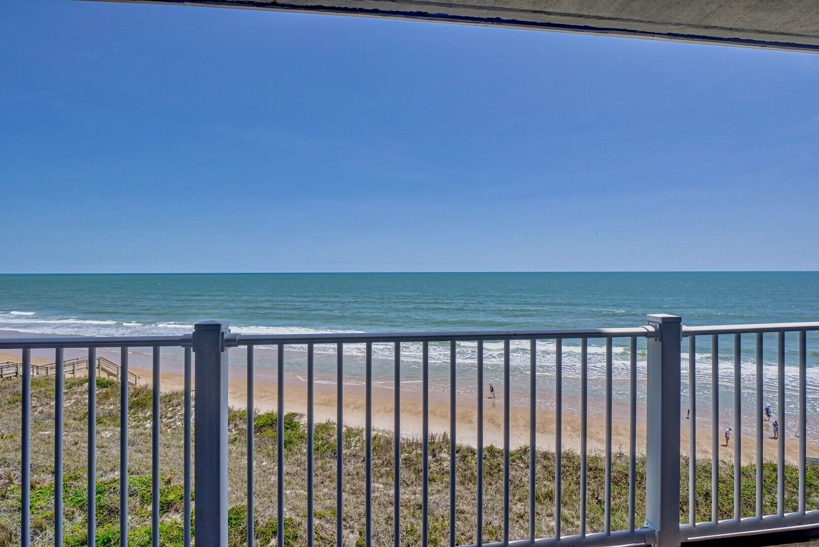 A beach view from a balcony with a railing, clear sky, and ocean.