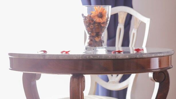 Et rundt træbord med glasplade, en vase med blomster og en stol med grøn pude.