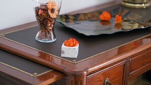 Et træskrivebord med en glaskrukke med visne blomster, en dekorativ bakke og en lille hvid æske med orange blomster.