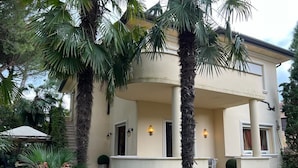 A two-story building with a curved balcony, palm trees, and a stone staircase leading to the entrance. - Villa MIMA with garden on two floors, 100 m from the sea. (Milano Marittima)