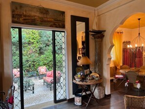 A room with a large glass door leading to an outdoor area with chairs and a table. There is a round table with various items on it, a tall column, and a chandelier.