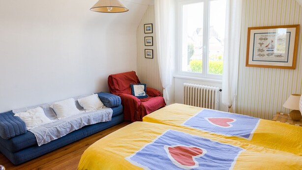 Une pièce avec un canapé bleu, un fauteuil rouge, deux lits avec une literie à motifs de cœur, une fenêtre avec des rideaux et un radiateur.