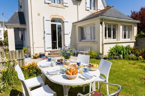 Une table à manger installée à l’extérieur, avec de la nourriture et des boissons, entourée de chaises, devant une maison dotée d’un jardin.