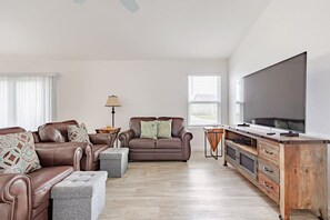 A living room with a brown leather sofa set, a grey ottoman, a wooden coffee table, a floor lamp, and a large flat-screen TV.
