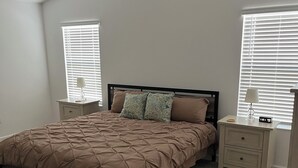 Une chambre à coucher comprenant un lit, deux tables de chevet, un ventilateur de plafond et deux fenêtres équipées de stores.