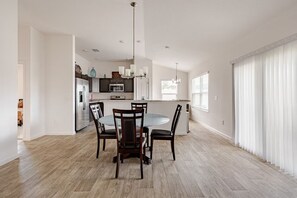 A spacious dining area with a round table and chairs, a kitchen in the background, and large windows with blinds.
