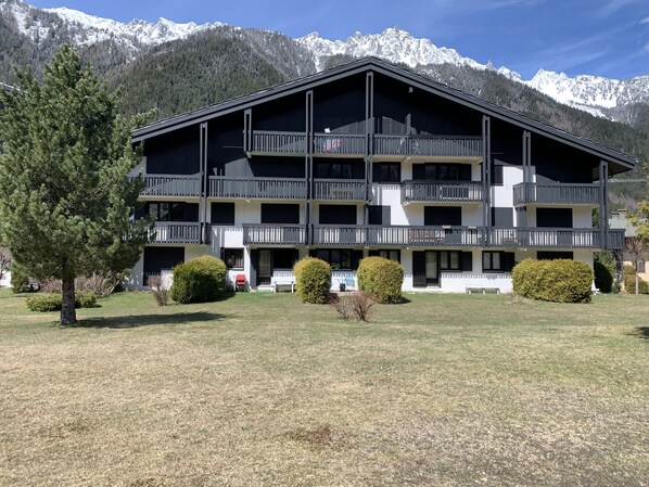 Un hotel de varios pisos con balcones, rodeado de un césped cuidado y árboles, con montañas nevadas al fondo.