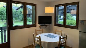 Zona de dinar amb taula i caires, una televisió sobre un suport i grans finestres amb vista a un barri residencial.