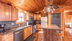 A kitchen with wooden cabinets, granite countertops, stainless steel appliances, and a wooden island with stools. - Elevated Farm Cabin | River Views & Access, Hot Tub & Thoughtful Finishes (Steelville)