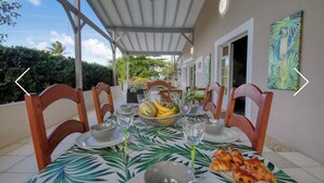 Uma área de refeições ao ar livre coberta, com uma mesa posta para dois, toalha de mesa com folhas tropicais, uma cesta de frutas e um prato de comida.