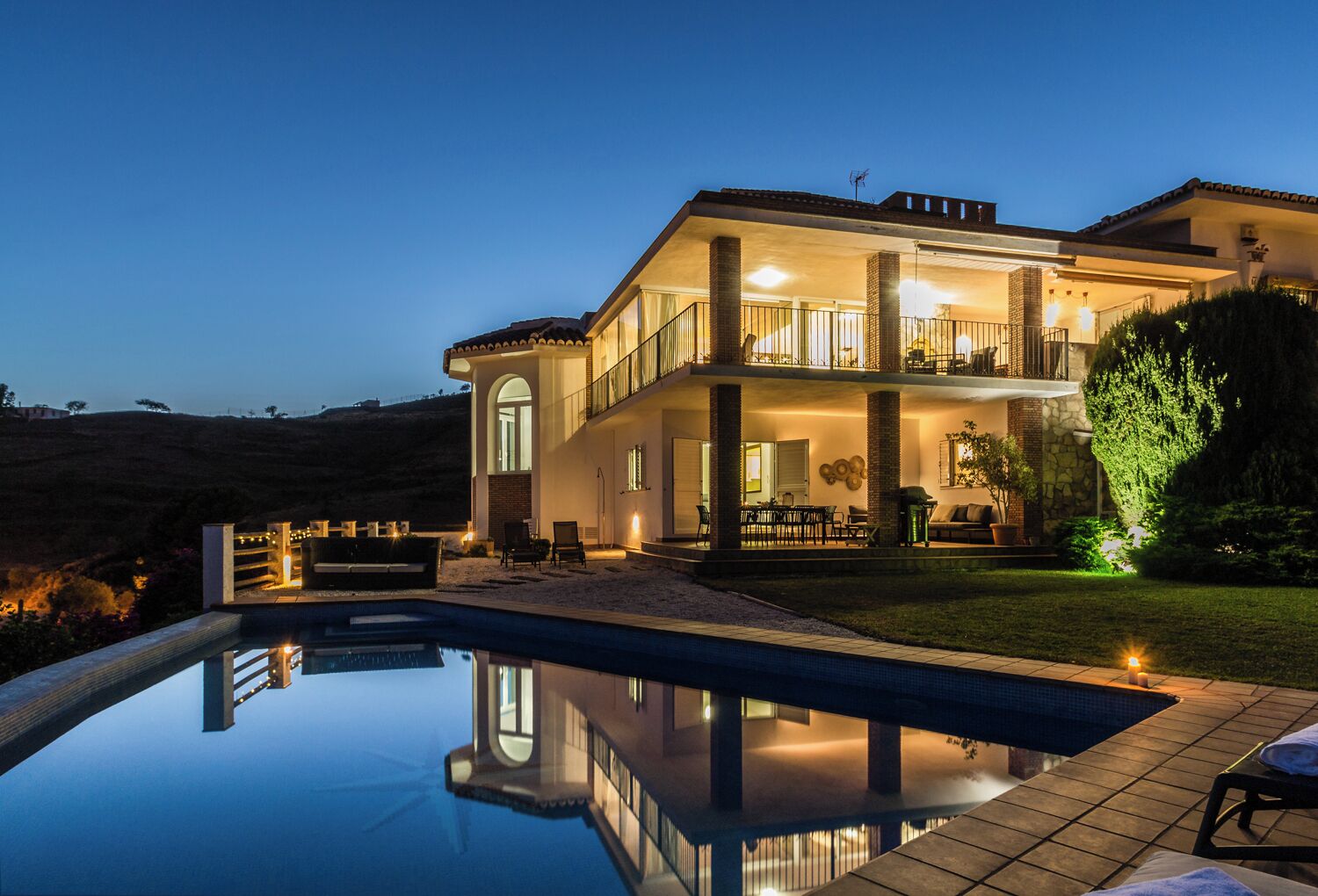 Una casa de dos pisos con piscina, mobiliario exterior y un balcón bien iluminado.