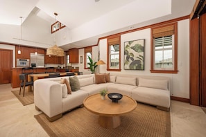 A spacious living area with a large L-shaped sectional sofa, a round wooden coffee table, and a kitchen in the background.