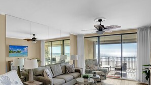 Ein Wohnzimmer mit einer Couch, zwei Lampen, einem Deckenventilator, einem Strandbild und einem Balkon mit Meerblick.