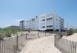 A multi-story hotel with a sandy beach path leading to the entrance, surrounded by a wooden fence.